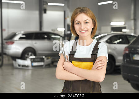 Handsome, charming woman smiling, looking at camera and holding tools, wrenches in hands. Female mechanic wearing in white t shirt and brown coveralls. Modern, professional autoservice. Stock Photo