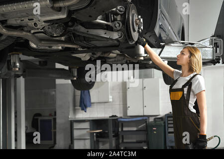 Beautiful young woman standing near lifted vehicle, observing and fixing brake discs with special tool. Girl wearing in white t shirt and coveralls, working in autoservice station. Stock Photo