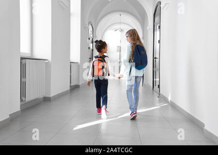 Two schoolgirls in jeans and sneakers with backpacks walking in long light hallway after lessons. Friends holding hands and talking in school. Stock Photo