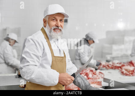 Elderly man working on butchery. Professional worker in white uniform, white cap, brown apron standing near table with meat, posing and looking at camera. Butcher holding knife and cutting. Stock Photo