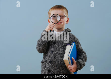 Cute little ginger boy in glasses looking through magnifying glass over blue Stock Photo