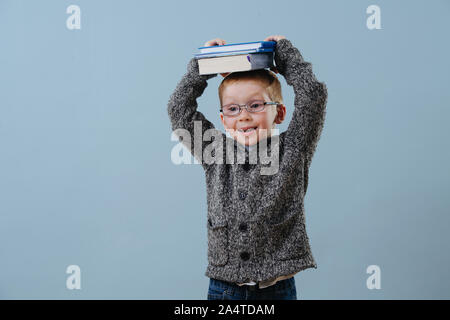 Cute little ginger boy in glasses holding pile of books on his head over blue Stock Photo