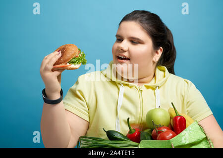 Chunky young brunette female is looking at hamburger with open mouse while keeping fruits and vegetables on her hand. Concept of temporal unhealthy temptation instead of choosing healthy eating. Stock Photo
