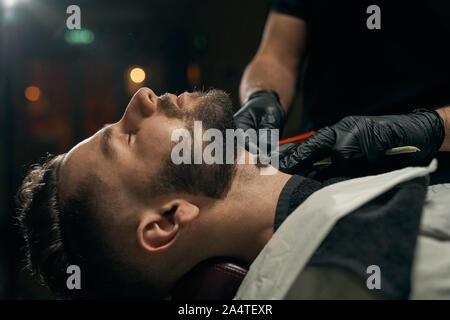 Side view of bearded handsome guy lying on couch while competent male barber in black gloves shaving his beard by sharp razor.Young client at barbershop is changing hairstyle and appearance Stock Photo