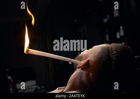 Close up of stylish man s head with white stick in his ear that burns in one end, sitting in armchair at modern barbershop. The process of ear hairs removal by burning fire Stock Photo