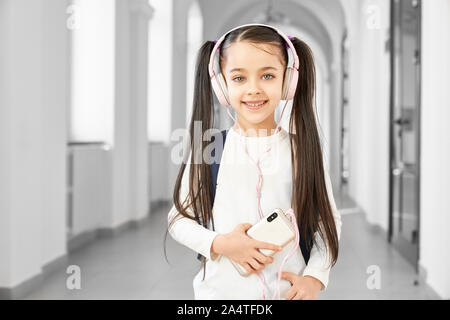 Portrait of pretty brunette school girl with funny hairstyle listening music on smart phone from big pink headphones. Smiling girl looking at camera, standing among corridor of beautiful school. Stock Photo