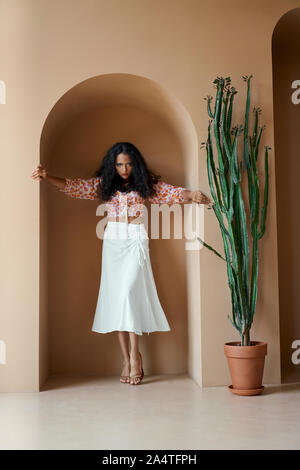Passionate beautiful young woman with tanned skin in trendy summer clothes standing in niche of wall near big green cactus. Female model with curly hair on high heels posing indoors Stock Photo
