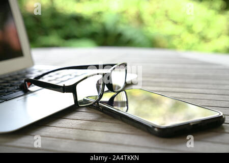 Laptop, cell phone and reading glasses on wooden table. Outdoor or green nature background. Stock Photo