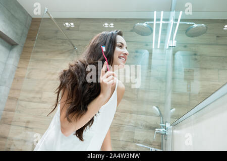 young beautiful woman standing in front of bathroom mirror while brushing her teeth Stock Photo