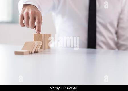 Business man interrupting domino effect by stopping wooden dominoes bricks from crumbling with his finger. Stock Photo