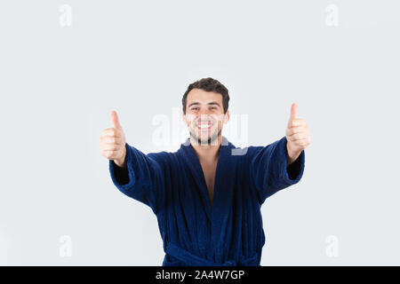 Portrait of cheerful young man in morning positive smiling showing thumbs up gesture, wears blue bathrobe, isolated over white background. Stock Photo
