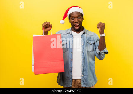 Portrait of amazed happy man in santa hat and casual denim shirt standing with raised hand showing yes gesture, holding bags, excited about shopping. Stock Photo