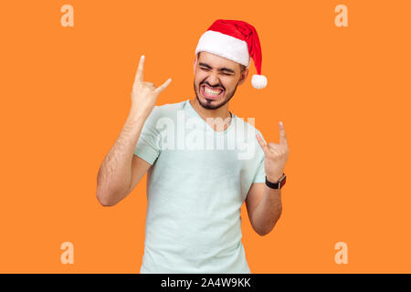 Portrait of extremely happy bearded young man in santa hat and casual white t-shirt standing with closed eyes, showing rock and roll hand gesture. ind Stock Photo