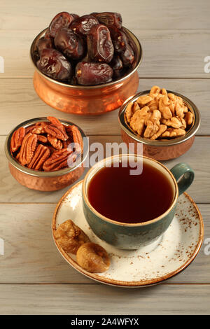 Vegetarian dessert: dried dates, pecans and walnuts in copper bowls, dried figs on a saucer and a cup of black tea on a gray wooden table. Closeup Stock Photo