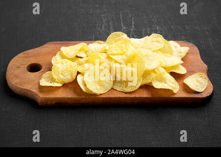 Crispy potato chips with salt on a rustic wooden board on a black surface, side view. Stock Photo
