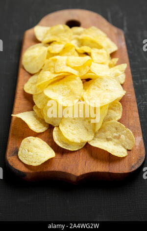 Crispy potato chips with salt on a rustic wooden board on a black surface, side view. Stock Photo