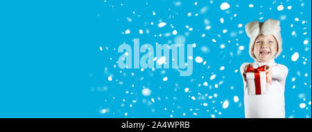 A cheerful boy in a white hare costume gives a gift with a red ribbon on a blue background. Snow is falling Stock Photo