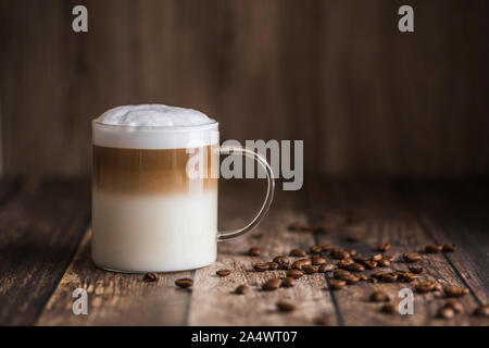 Cafe latte macchiato layered coffee in a see through glass coffee cup. The cup is on a wooden background with coffee beans on the table next to the cu Stock Photo