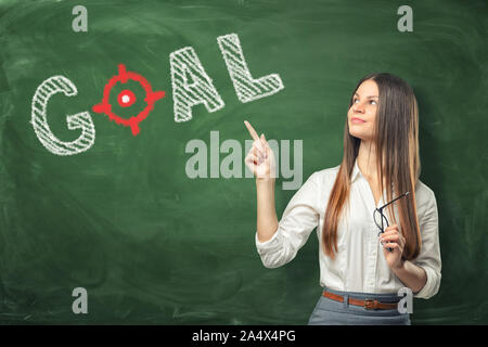 Young woman holding her glasses and pointing finger at word 'goal' written on green chalkboard wall. Stock Photo
