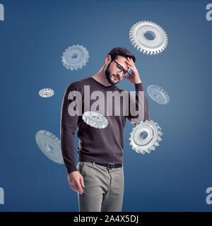 A man in glasses stands deep in thought while being surrounded by several large metal spur gears. Stock Photo