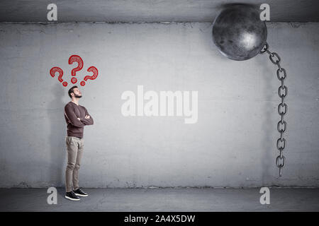 A bearded man stands with large red question signs above him and looks at a wrecking ball stuck to the ceiling. Stock Photo