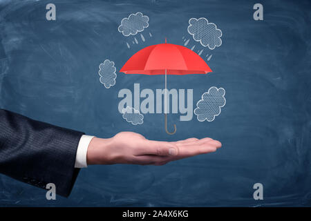A businessman's hand turned up and a small red umbrella hovering above it on chalkboard background. Stock Photo