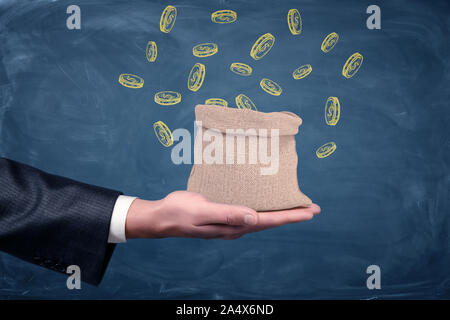 A businessman's hand turned up and a small money bag standing on it with drawn coins on blue background. Stock Photo