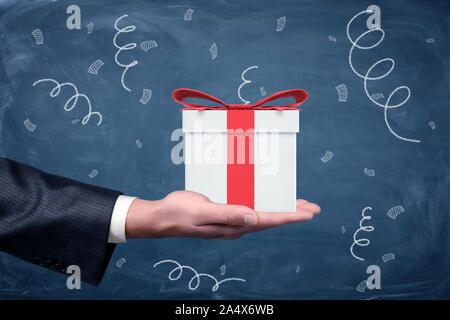 A businessman's hand turned up and a small gift box with a red bow standing on chalkboard background. Stock Photo