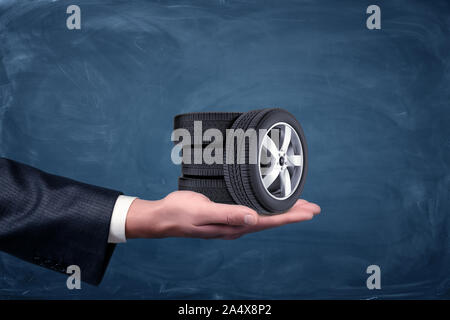 A businessman's hand on blue chalkboard background holding four tiny car wheels. Stock Photo