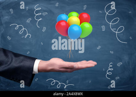 A businessman's hand turned up with a set of colorful balloons tied together hovering above it on chalkboard background. Stock Photo