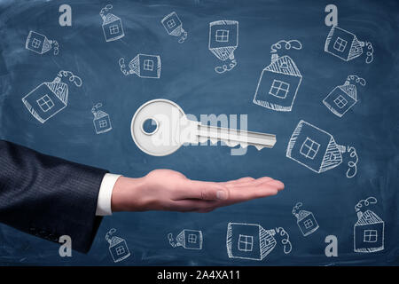 A businessman's hand turned palm up with a giant key hovering above it and house doodles on a blackboard background. Stock Photo