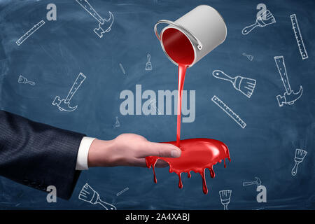 A businessman's hand turned palm up receives a handful of red paint flowing from a small overturned bucket. Stock Photo