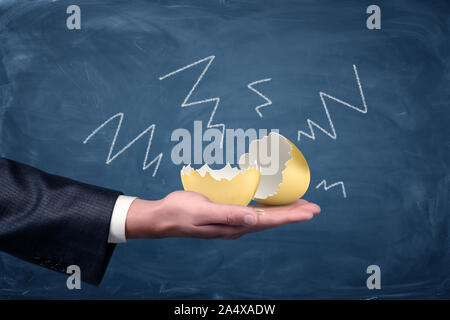A large cracked and empty golden egg lying on a businessman's palm with negative arrows drawn on a chalkboard around it. Stock Photo