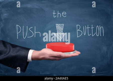 A large red push button resting on businessman's palm under a chalkboard drawing saying Push the Button. Stock Photo