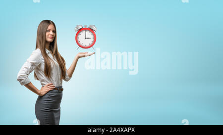 A blond businesswoman stands and holds one hand palm up with red retro alarm clock hovering above it. Stock Photo