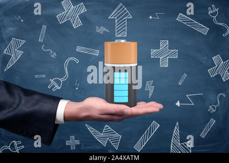 A male businessman's hand holds up a large fully-charged battery on a blue blackboard background. Stock Photo