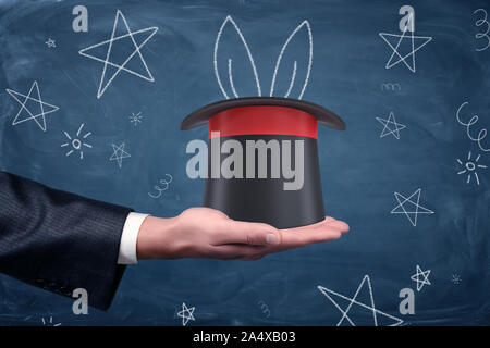 A businessman's palm with a magician's hat resting on it and drawings of stars and rabbit ears around. Stock Photo