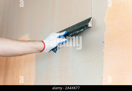 A worker is applying putty on a fiberglass mesh on the wall. He is using a wide spatula. Stock Photo