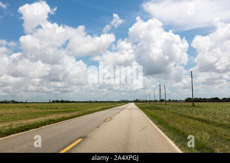 White clouds and blue sky over an emtpy two lane highway stretches to the horizon with empty fields on either side in the Everglades, Homestead, Florida
