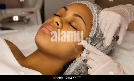Aesthetic corrective treatments concept. Cosmetology. Facelift in spa salon. Beautician makes injections afro american girl in cheekbone. Smoothing of Stock Photo