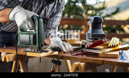 Adult carpenter craftsman wears protective leather gloves, with electric saw working on cutting a wooden table. Construction industry, housework do it Stock Photo