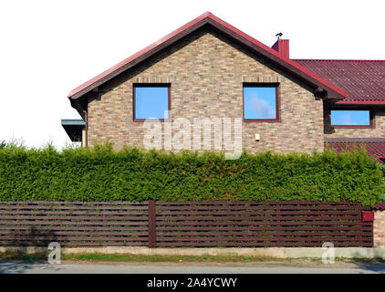 Behind a wooden fence and green coniferous hedge  is the side wall of a  standard no name yellow rural brick house. Blue sky reflections in windows. I Stock Photo