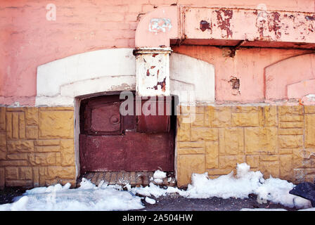 Yellow, pink colors wall with old red boarded up window on the ground level, ventilation pipe, melting snow on asphalt, grunge horizontal city detail Stock Photo