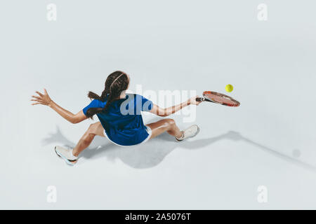 Young woman in blue shirt playing tennis. She hits the ball with a racket. Indoor studio shot isolated on white. Youth, flexibility, power and energy. Negative space. Top view. Stock Photo