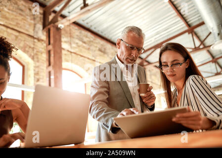 Sharing experience. Senior man in formal wear holding coffee cup and explaining something to his young female colleague while working together in the Stock Photo