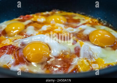 Freshly cooked appetizing fried eggs with red sweet tomatoes in a hot pan. Delicious homemade food. Four yolks of chicken eggs. Stock Photo
