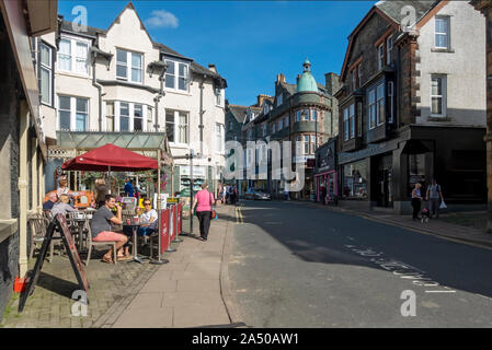 People sitting drinking sat outside cafe in the town centre shops stores in summer Keswick Cumbria England UK United Kingdom GB Great Britain Stock Photo