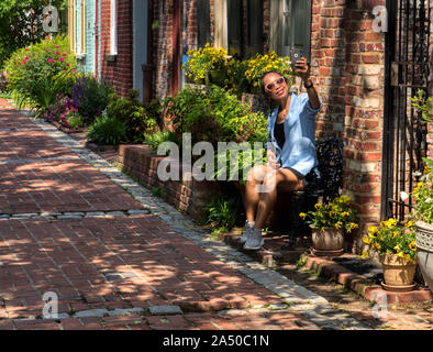 Asian woman wearing sunglasses sitting on chair surrounded by plants taking selfie in front of brick building Stock Photo