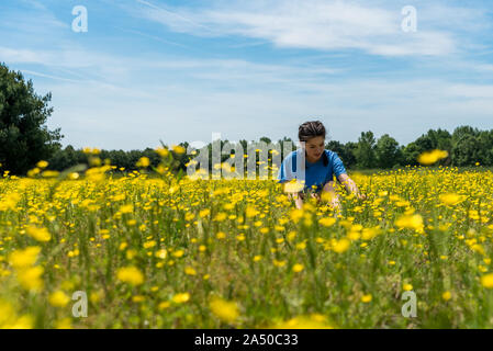 Low angle view of teen girl with braided hair sitting in large field with yellow flowers and trees in the background Stock Photo
