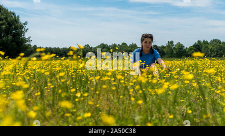 Low angle view of teen girl sitting in large field with yellow flowers and trees in the background Stock Photo